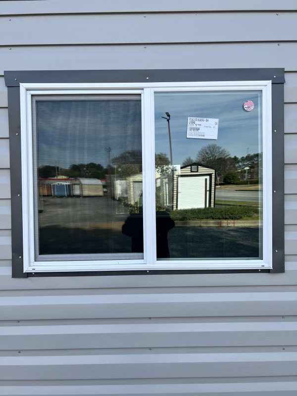 48×36-2-Light-Slider-600×800 Window view of a portable storage building showcasing customizable features, with a reflection of nearby structures and a price tag visible on the glass.