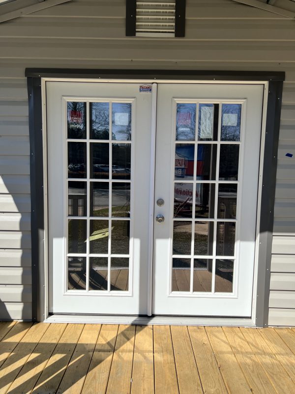 60×68-15-Light-French-Door-600×800 Double light French doors with grid design, leading to a portable storage building, featuring a wooden deck and metal siding.