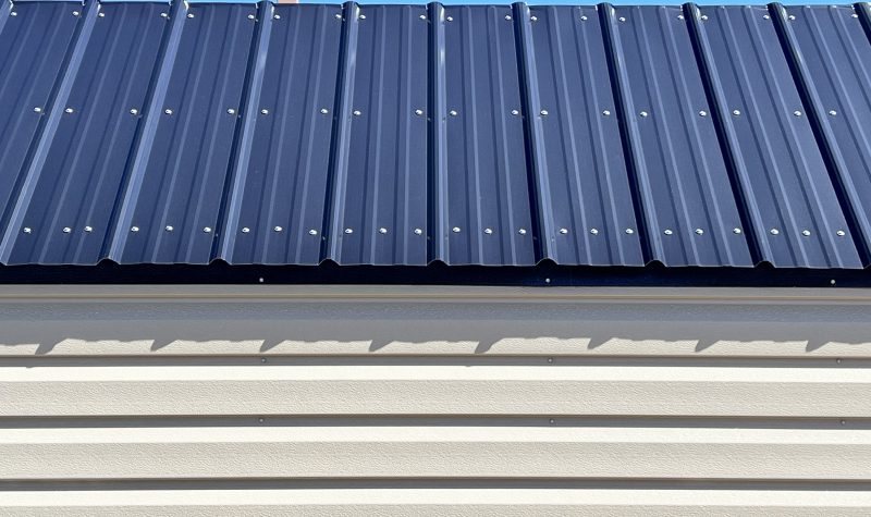 Colored-Steel-Roof-800×475 Close-up of a blue metal roof with visible screws above light-colored siding, showcasing customizable roofing options for portable buildings and sheds.