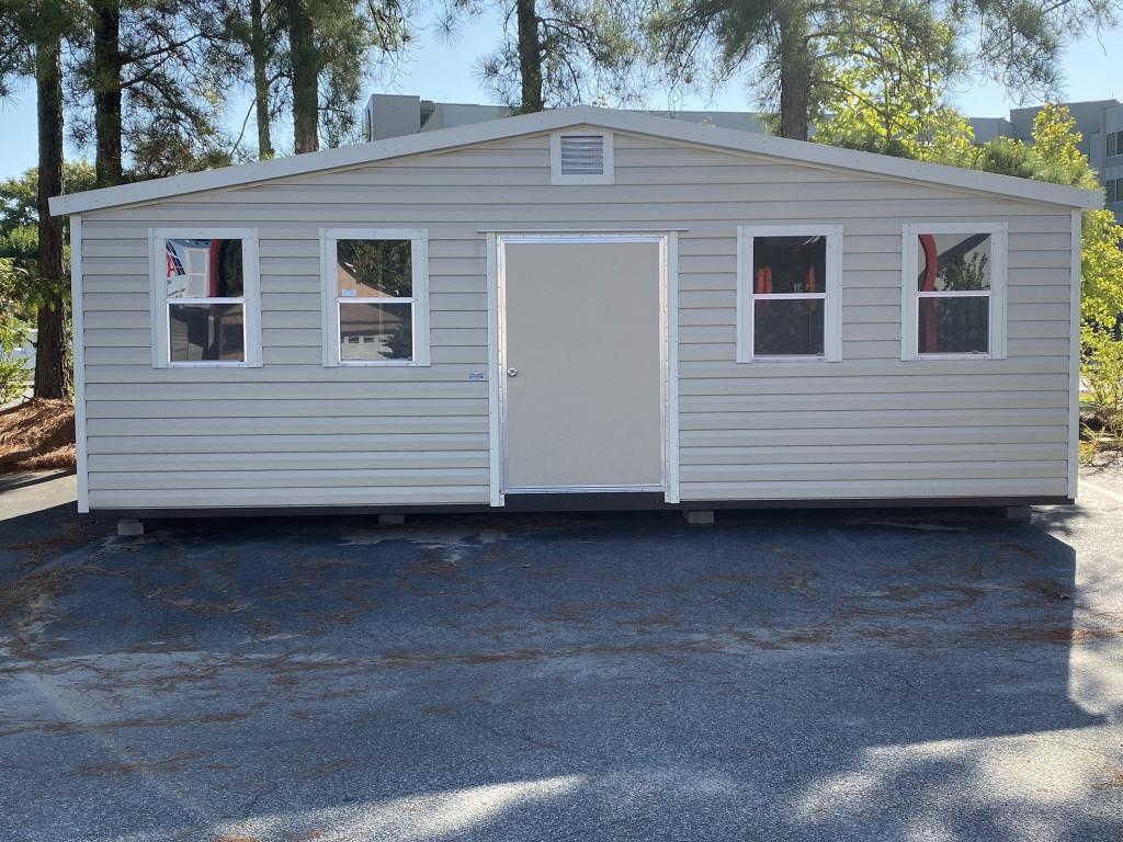 Side gable portable cabin with a sloped roof, front door, and multiple windows, set against a backdrop of trees, showcasing functional design for storage or workspace.