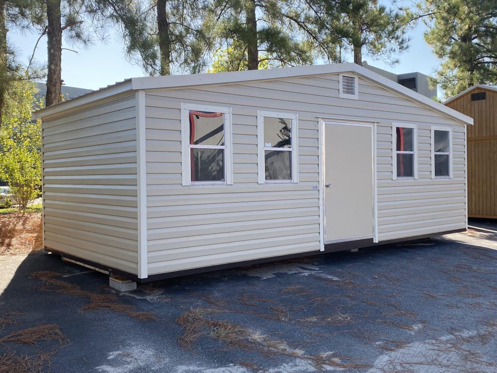 Side gable portable cabin with beige siding, multiple windows, and a front door, situated in a shaded outdoor environment, showcasing functional design for storage or workspace needs.