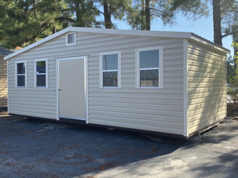 Side-gable portable cabin with beige siding, front door, and multiple windows, set in a landscaped outdoor area, showcasing its functional design for storage or workspace.
