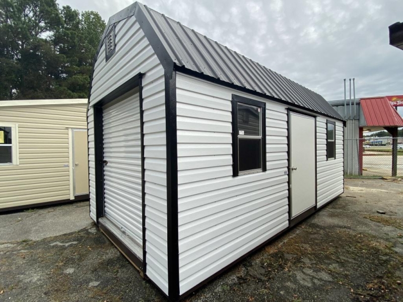 Portable-Lofted-Mini-Garages-Albany-GA-e1574150597890 Portable lofted mini garage with a gambrel roof design, featuring a white exterior and black trim, positioned outdoors among other storage structures.